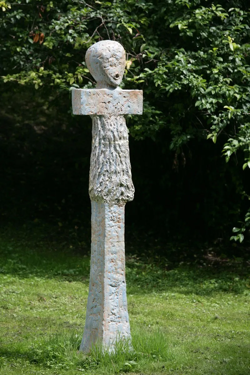 Tall pale figure with a head above a crossbar in a garden