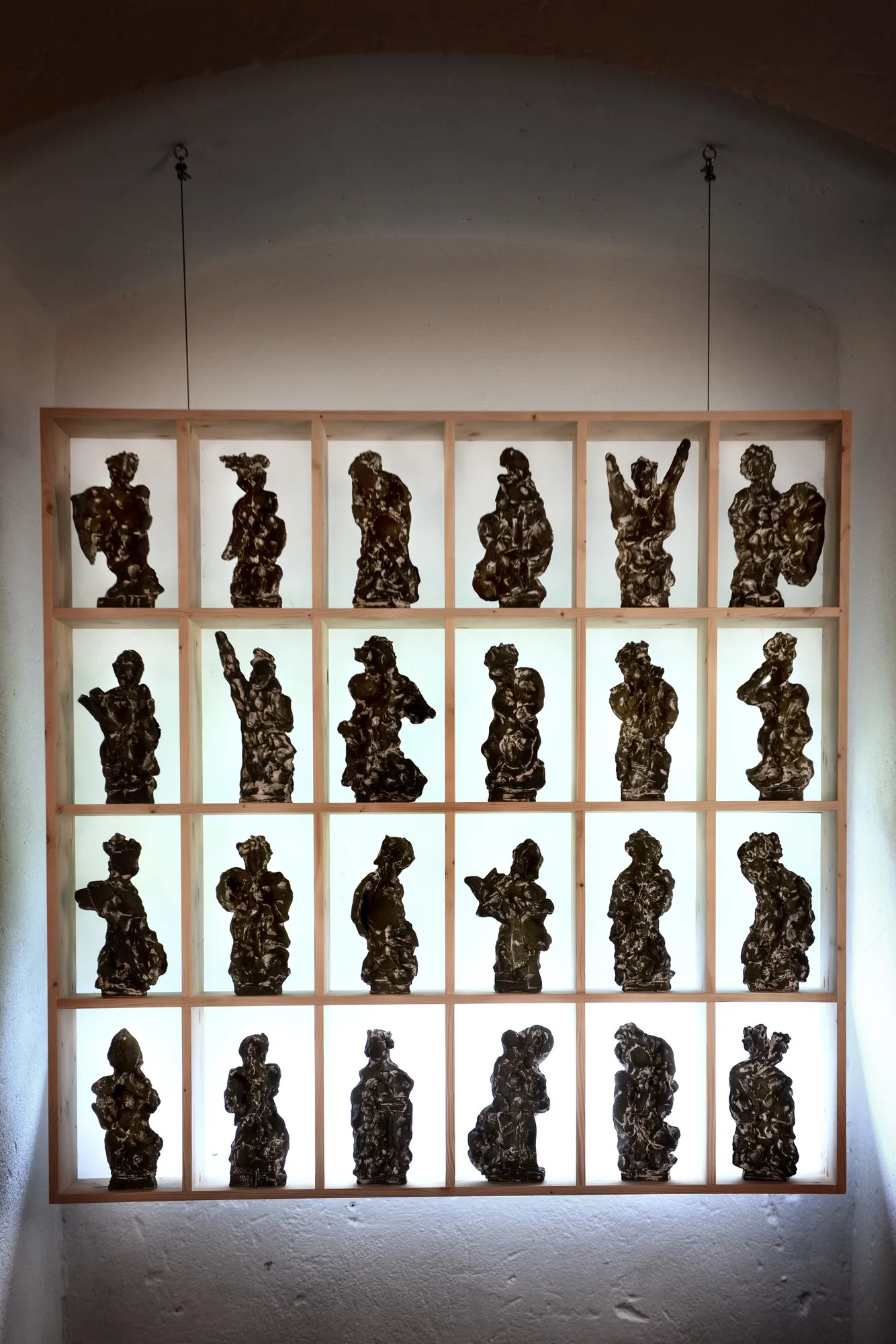 Dozens of small dark figures in a grid of pale wooden shelves against a window