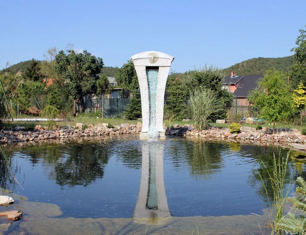 White gate-shaped sculpture with a jet of water, reflected in a garden pond