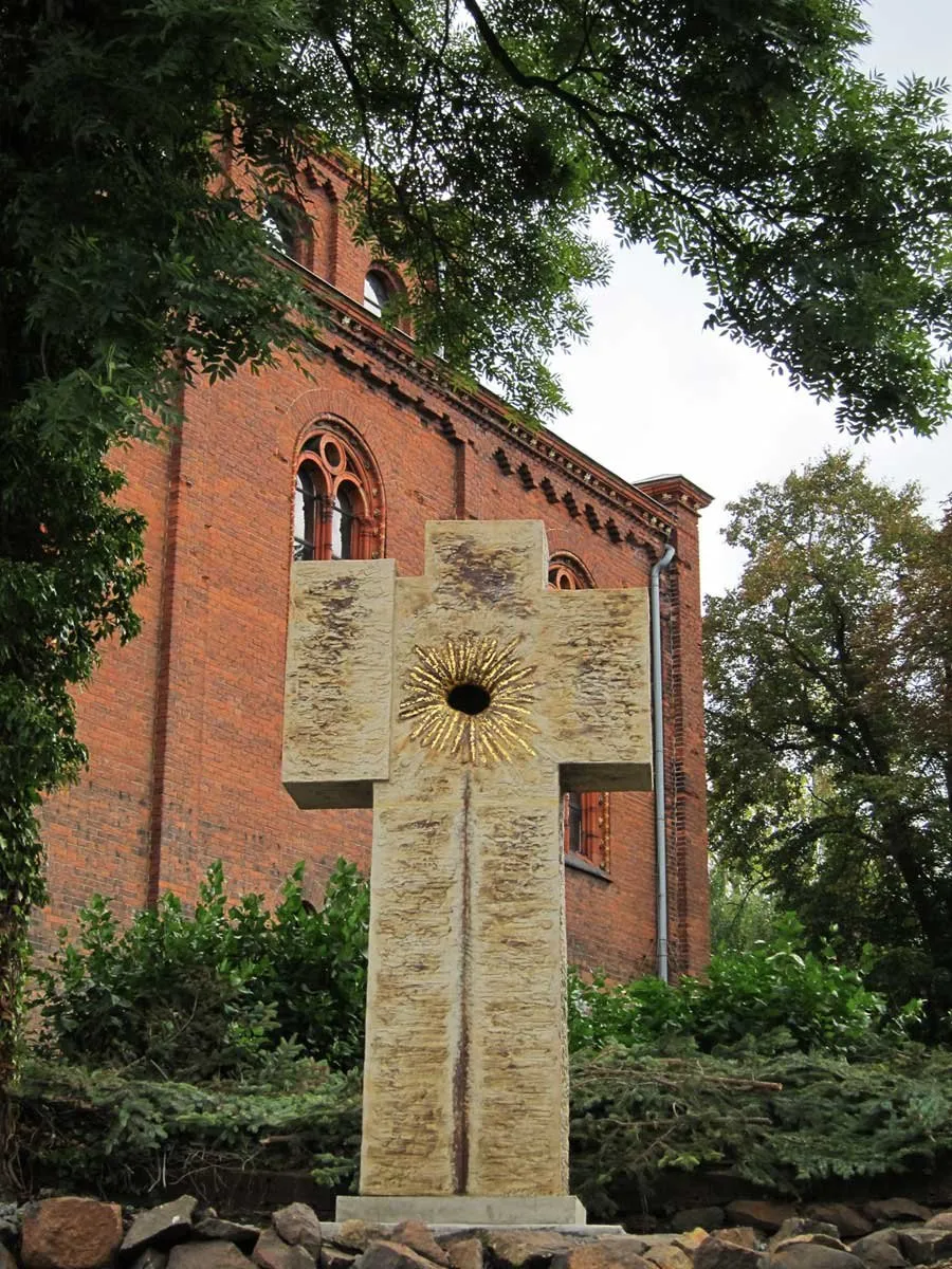 Světlý keramický kříž se zlatým paprsčitým středem před cihlovým kostelem
