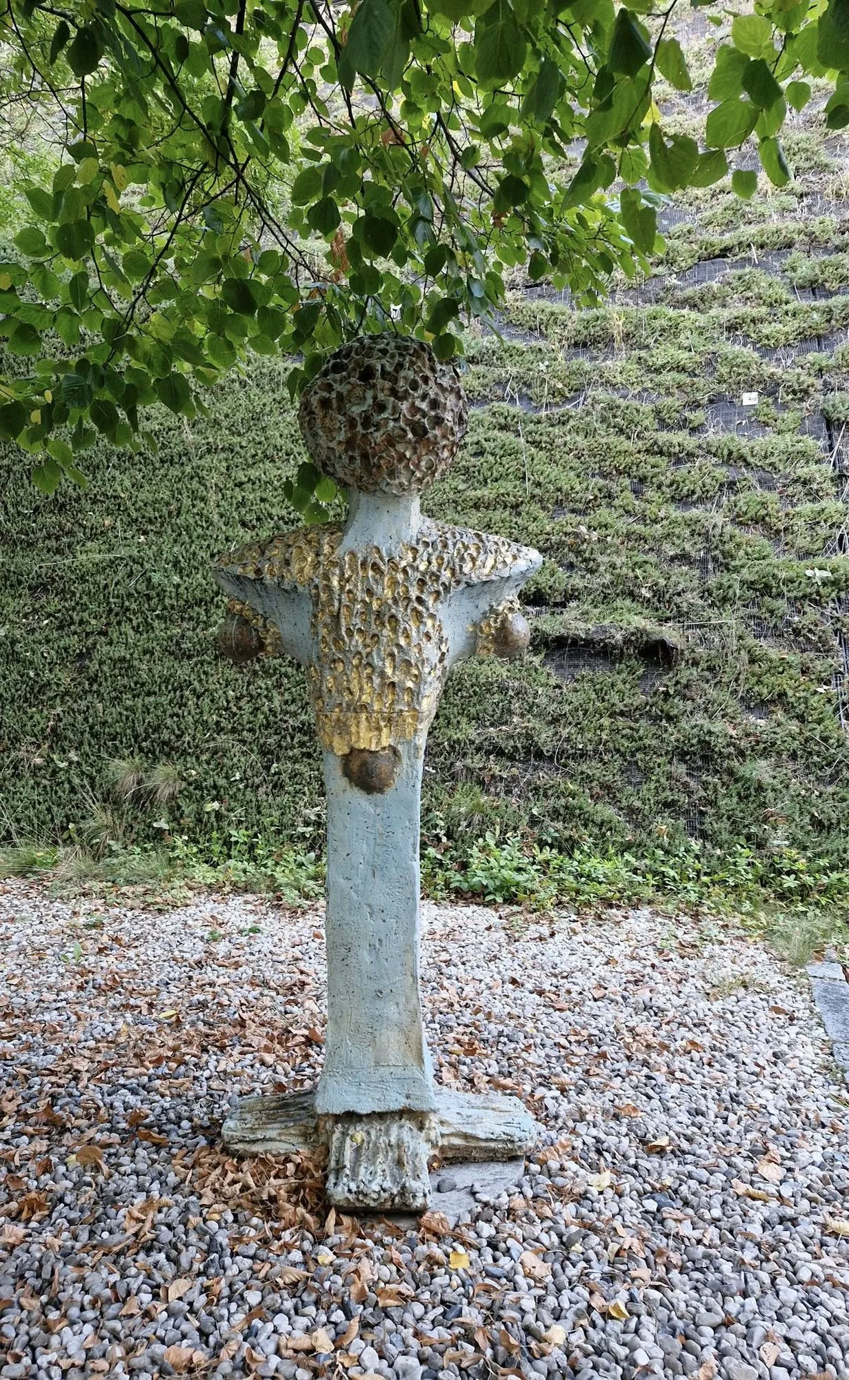 Blue figure with a textured head in a gravel garden under a tree
